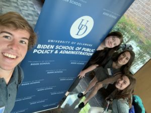 Four students smile together, standing around a University of Delaware Biden School of Public Policy & Administration sign