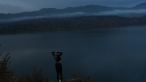 A woman wearing exercise clothes stands facing away from the camera, looking over a large lake with foggy mountains in the background.