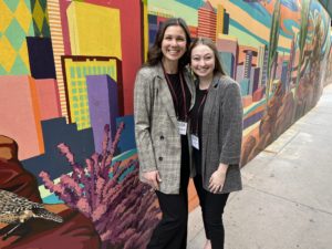 Two women in professional attire smiling in front of a colorful wall mural