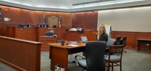 Photo of Leslie Schenk, a young woman with long, blonde hair wearing a dark suit and smiling while seated in an otherwise empty courtroom