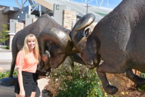 Photo of Leslie Schenk, a young woman with long, blonde hair dressed professionally and smiling next to a large, bronze statue of two rams butting heads outside the football stadium on CSU's campus