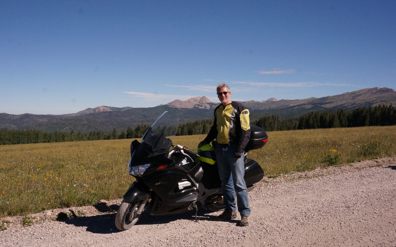 Professor Mike Palmquist stands beside his motorcycle on the side of a dirt path with mountains in the background.