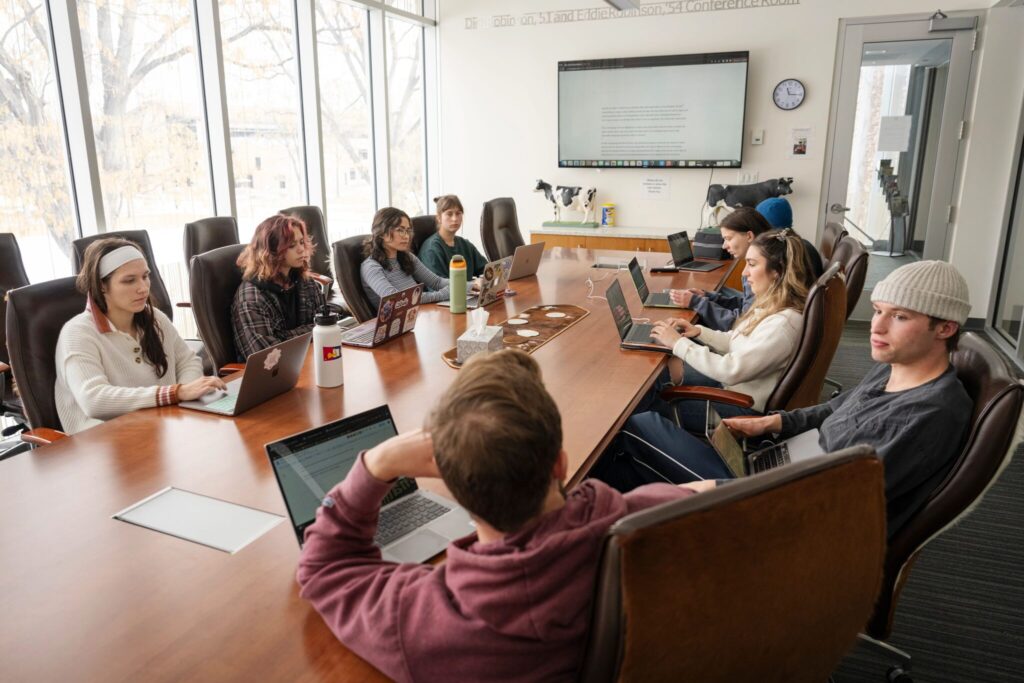 Classroom of students sitting around a conference table, looking at each other and laptops